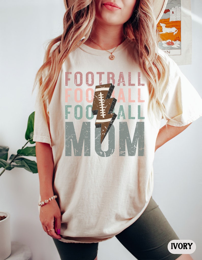 Op de afbeelding: Een cr&egrave;mekleurig T-shirt met een verweerde grafische afbeelding waarop "Football Mom" staat met een voetbal- en bliksemontwerp. De tekst is in tinten roze, groen en grijs.