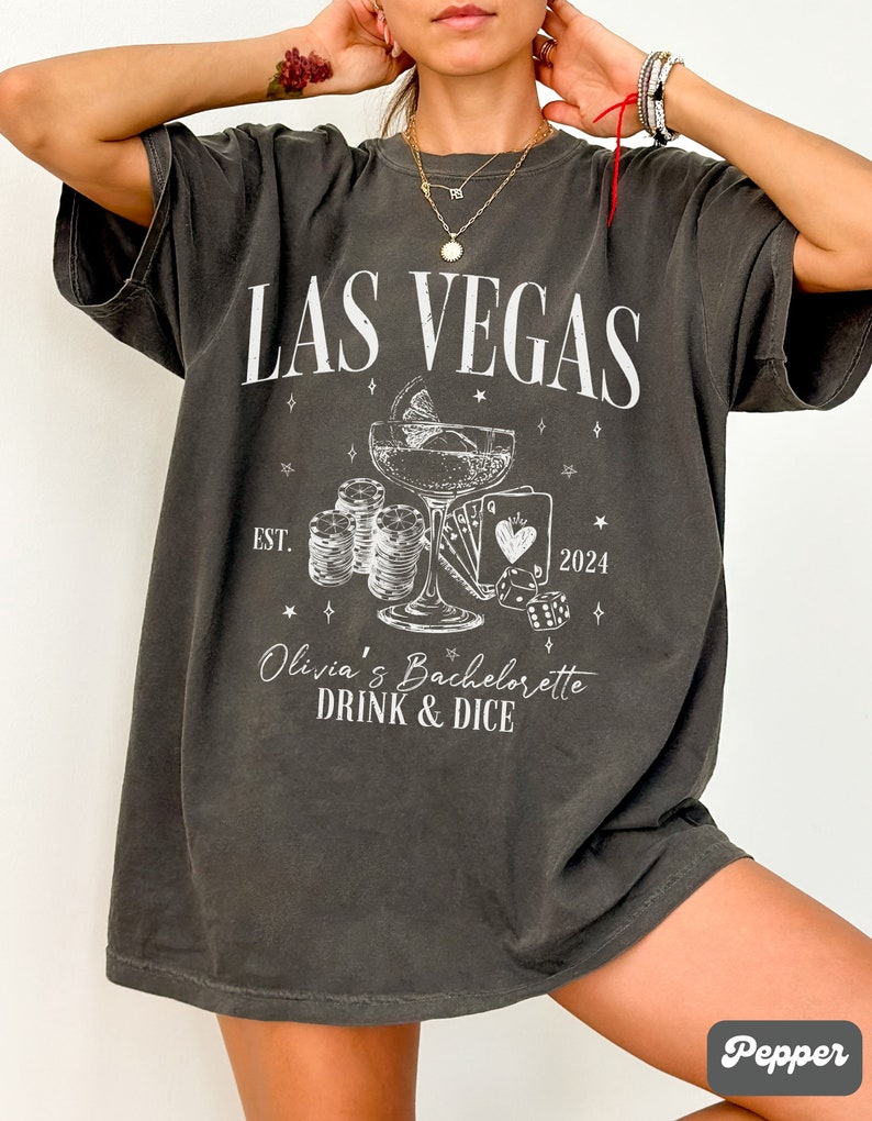 May include: A dark gray t-shirt with a white graphic design that says "Las Vegas" with a cocktail glass, playing cards, dice, and the text "Olivia's Bachelorette Drink & Dice" and "Est. 2024".