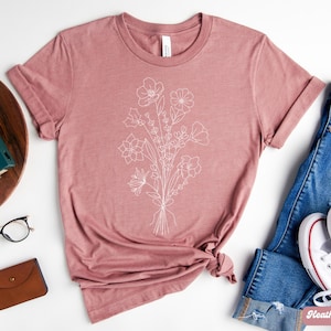 K&ouml;nnte beinhalten: Ein T-Shirt in Heide-Mauve mit einer wei&szlig;en Linienzeichnung eines Blumenstrau&szlig;es. Das Shirt wird auf einem wei&szlig;en Hintergrund mit einer Jeans, einer Kamera und einer Brille gezeigt.