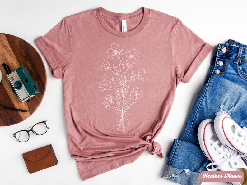 K&ouml;nnte beinhalten: Ein T-Shirt in Heide-Mauve mit einer wei&szlig;en Linienzeichnung eines Blumenstrau&szlig;es. Das Shirt wird auf einem wei&szlig;en Hintergrund mit einer Jeans, einer Kamera und einer Brille gezeigt.