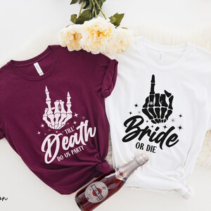 Puede incluir: Dos camisetas con un dise&ntilde;o de mano de esqueleto. La camiseta granate dice "Till Death Do Us Party" y la camiseta blanca dice "Bride or Die".