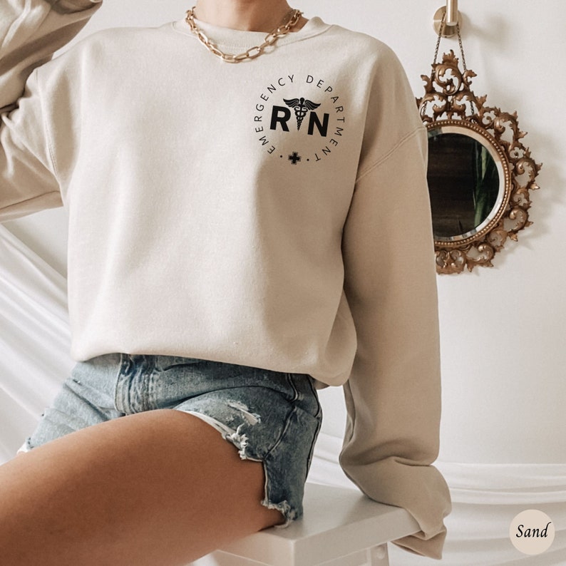 Peut inclure: Un sweat-shirt beige avec un graphique noir et blanc d'un caduc&eacute;e m&eacute;dical et le texte "Emergency Department RN".