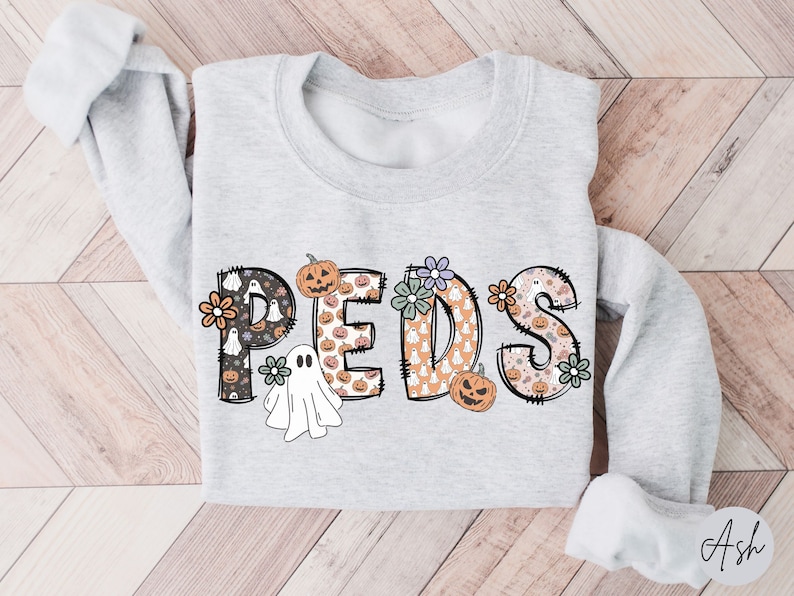May include: A light gray sweatshirt with the word "PEDS" in a Halloween-themed design. The letters are decorated with pumpkins, ghosts, and floral accents. The sweatshirt has long sleeves.