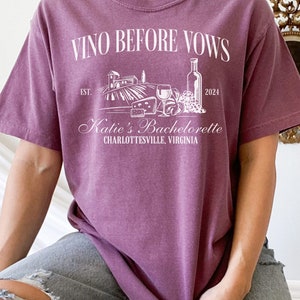 May include: A purple t-shirt with the text "Vino Before Vows" and a graphic of a vineyard, wine bottle, grapes, and cheese. The text "Katie's Bachelorette Charlottesville, Virginia" is also on the shirt.