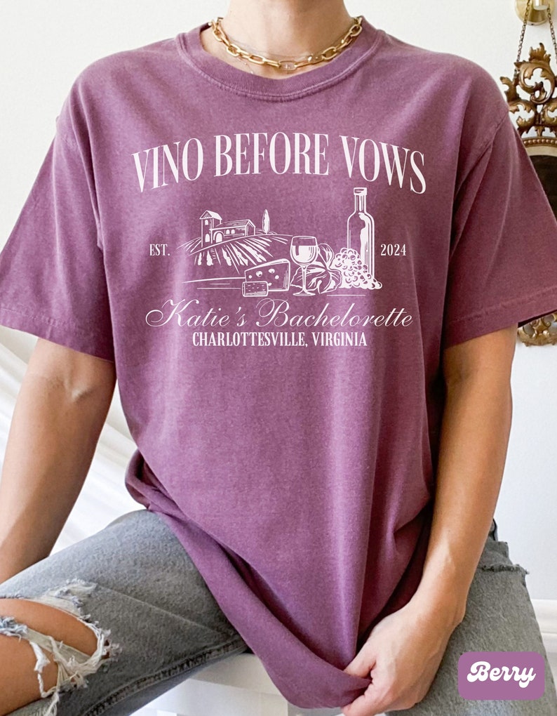 May include: A purple t-shirt with the text "Vino Before Vows" and a graphic of a vineyard, wine bottle, grapes, and cheese. The text "Katie's Bachelorette Charlottesville, Virginia" is also on the shirt.