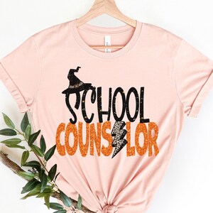 May include: A pink t-shirt with the text "SCHOOL COUNSELOR" in black and orange glitter. A witch's hat with a black cat is above the word "SCHOOL".