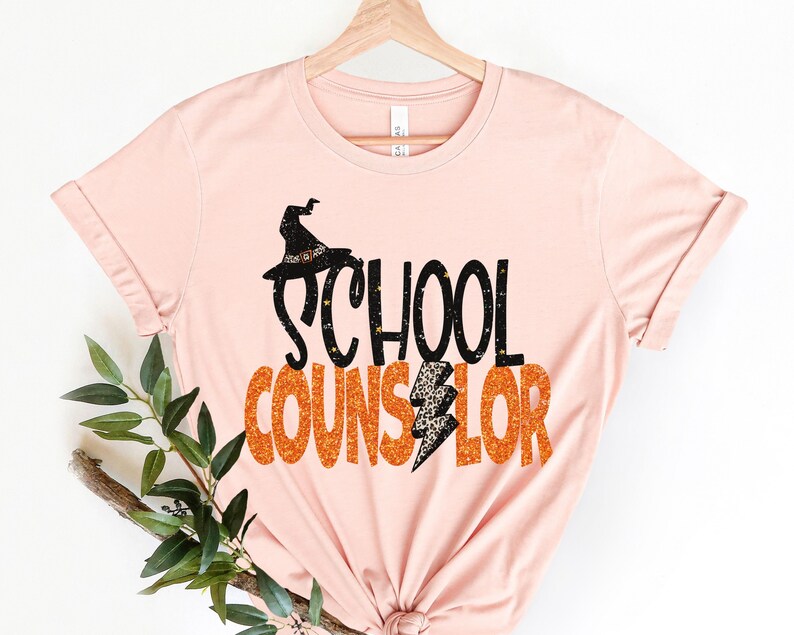 May include: A pink t-shirt with the text "SCHOOL COUNSELOR" in black and orange glitter. A witch's hat with a black cat is above the word "SCHOOL".
