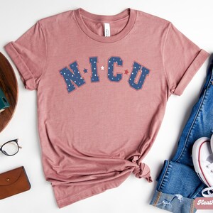 May include: A heather mauve t-shirt with the text "NICU" in blue and red letters with white stars. The shirt is made of a soft, comfortable material and is perfect for everyday wear.