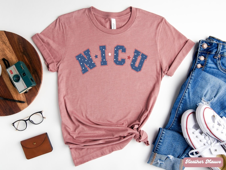 May include: A heather mauve t-shirt with the text "NICU" in blue and red letters with white stars. The shirt is made of a soft, comfortable material and is perfect for everyday wear.