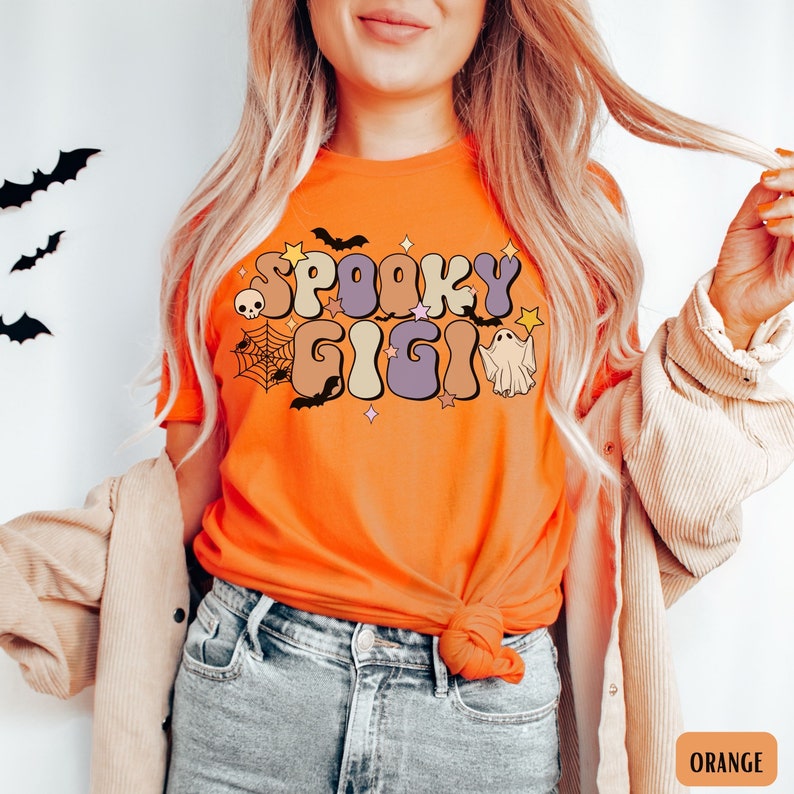 May include: Orange t-shirt with the text "Spooky Gigi" in a retro font. The text is surrounded by bats, stars, a spider web, a skull, and a ghost.