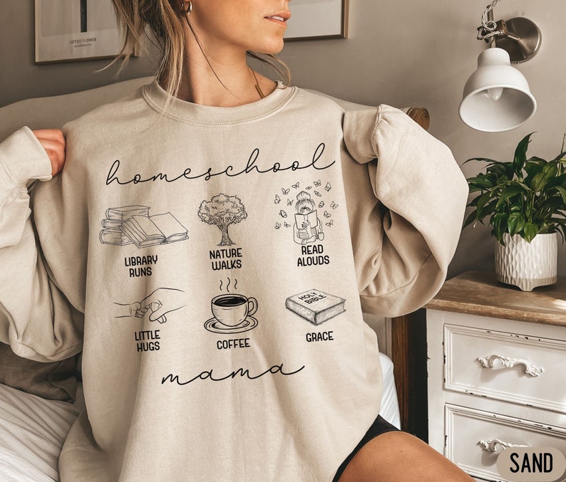 Puede incluir: Una sudadera beige con el texto "homeschool mama" e ilustraciones de libros, un &aacute;rbol, una taza de caf&eacute;, una mano sosteniendo una mano peque&ntilde;a, una persona leyendo un libro y un libro con el texto "Holy Bible" y la palabra "grace" debajo.