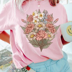Pode incluir: Um moletom rosa claro com um gr&aacute;fico de buqu&ecirc; floral detalhado em tons de rosa, branco, amarelo e azul. A palavra "Blossom" est&aacute; impressa no canto inferior direito. A pessoa est&aacute; usando shorts azuis claros e um colar.