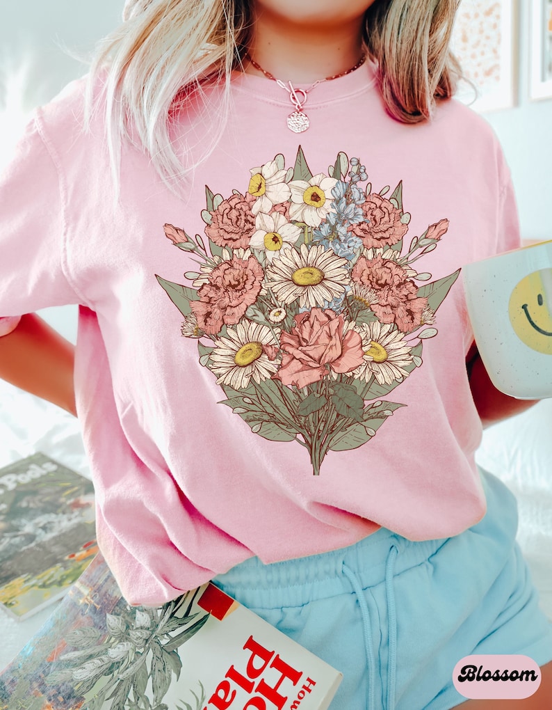 Pode incluir: Um moletom rosa claro com um gr&aacute;fico de buqu&ecirc; floral detalhado em tons de rosa, branco, amarelo e azul. A palavra "Blossom" est&aacute; impressa no canto inferior direito. A pessoa est&aacute; usando shorts azuis claros e um colar.