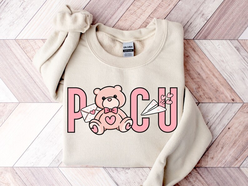 Peut inclure: Un sweat-shirt beige clair avec un motif graphique rose qui dit "POCU" avec un ours en peluche tenant une lettre et un avion en papier avec des c&oelig;urs au-dessus.