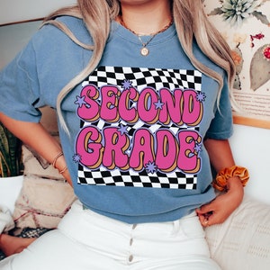 以下が含まれることがあります： チェック柄の背景に「Second Grade」というピンクとパープルの文字が書かれた、ブルーのジーンズ地のTシャツです。