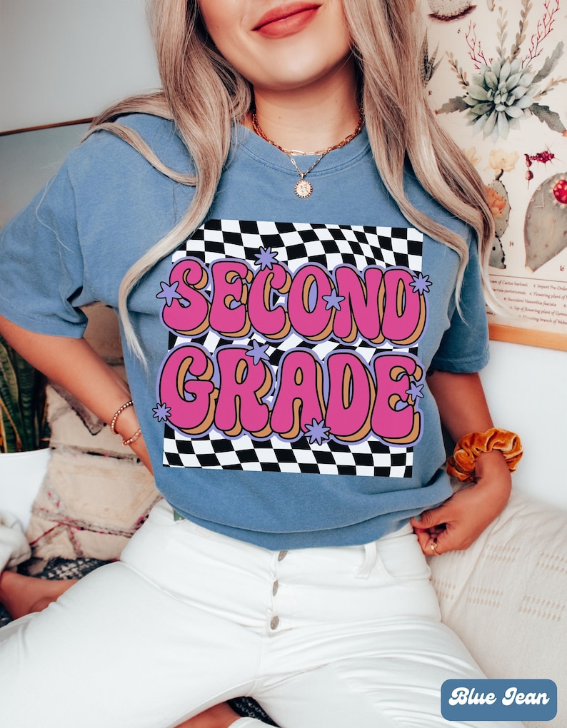 以下が含まれることがあります： チェック柄の背景に「Second Grade」というピンクとパープルの文字が書かれた、ブルーのジーンズ地のTシャツです。