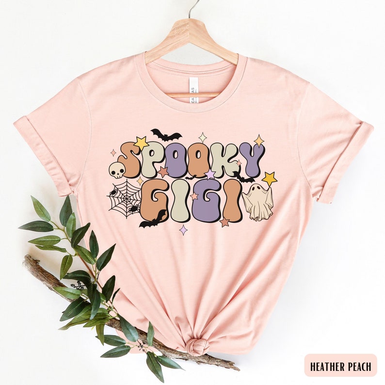 May include: A pink t-shirt featuring a retro-style graphic that reads "Spooky Gigi" in colourful letters. The graphic also includes bats, stars, a ghost, and a spider's web.