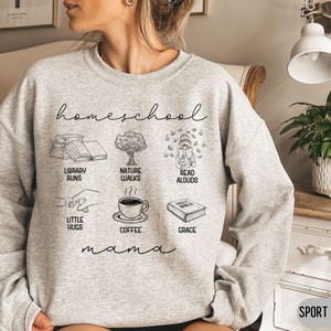 Puede incluir: Una sudadera gris con el texto "homeschool mama" e ilustraciones de libros, un &aacute;rbol, una persona leyendo, caf&eacute; y una biblia. Tambi&eacute;n se incluye el texto "library runs", "nature walks", "read aloud", "little hugs", "coffee" y "grace".