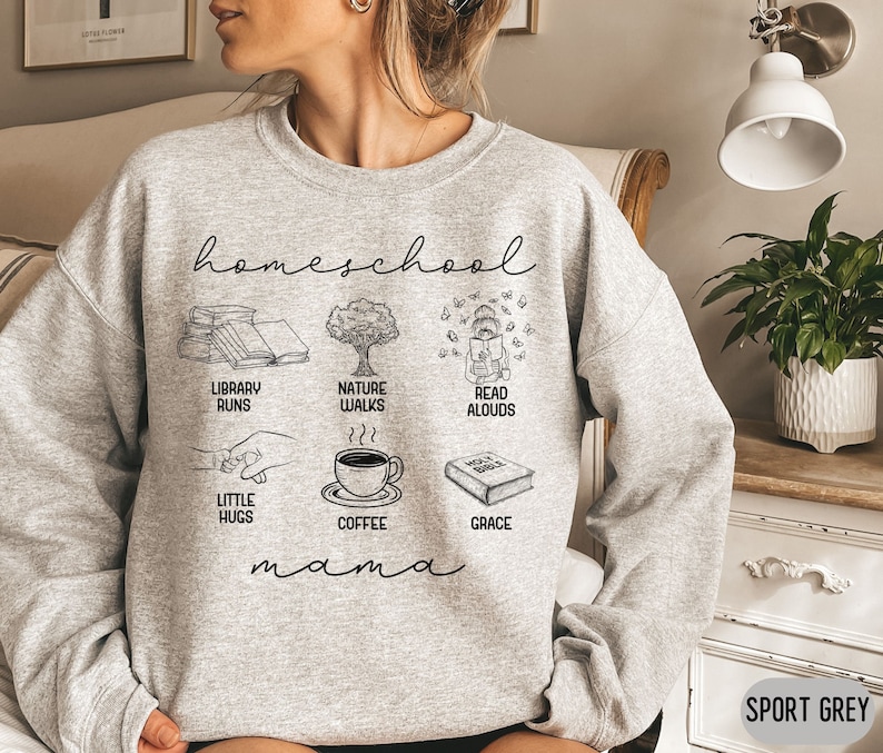 Puede incluir: Una sudadera gris con el texto "homeschool mama" e ilustraciones de libros, un &aacute;rbol, una persona leyendo, caf&eacute; y una biblia. Tambi&eacute;n se incluye el texto "library runs", "nature walks", "read aloud", "little hugs", "coffee" y "grace".