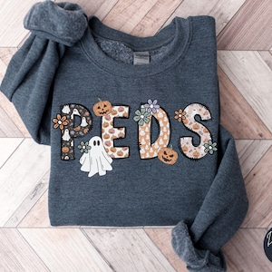May include: Dark grey heather sweatshirt with the word "PEDS" in a Halloween-themed design. The letters are decorated with pumpkins, ghosts, and flowers. The sweatshirt is folded on a wooden surface.