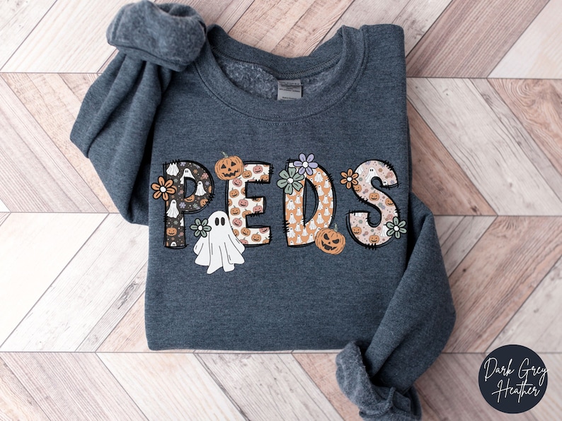May include: Dark grey heather sweatshirt with the word "PEDS" in a Halloween-themed design. The letters are decorated with pumpkins, ghosts, and flowers. The sweatshirt is folded on a wooden surface.