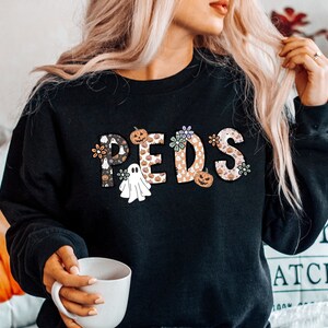 May include: Black sweatshirt with the word "PEDS" in a decorative font. The letters are filled with Halloween-themed patterns, including pumpkins, ghosts, and flowers. The sweatshirt is a crew neck style.