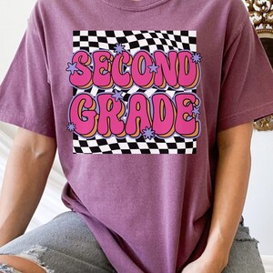 以下が含まれることがあります： チェック柄の背景に、ピンクとオレンジの文字で「Second Grade」と書かれたレトロなグラフィックがプリントされたパープルのTシャツです。