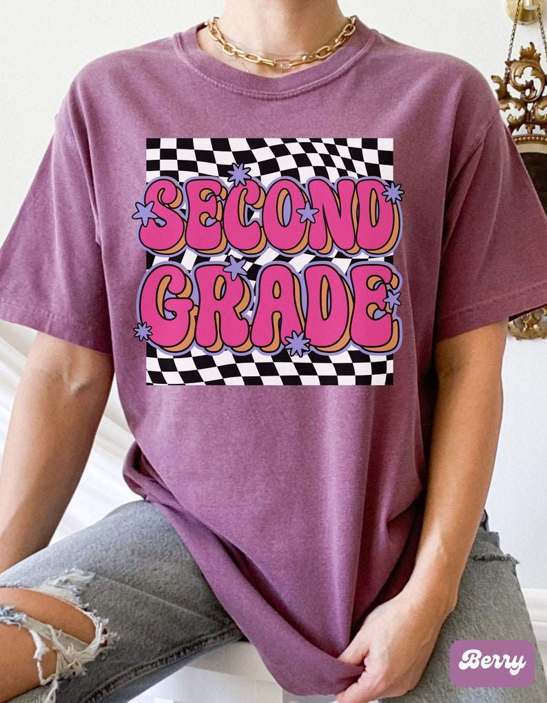 以下が含まれることがあります： チェック柄の背景に、ピンクとオレンジの文字で「Second Grade」と書かれたレトロなグラフィックがプリントされたパープルのTシャツです。