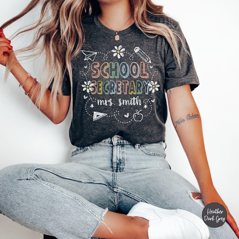 Peut inclure: T-shirt gris fonc&eacute; chin&eacute; avec l'inscription "SCHOOL SECRETARY mrs. smith" dans un design color&eacute; et fantaisiste. Le motif comprend un avion en papier, un crayon, des fleurs, une r&egrave;gle, une pomme et des c&oelig;urs.