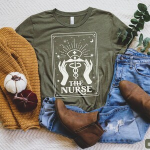 May include: A green t-shirt featuring a white graphic of a tarot card. The card depicts a white caduceus symbol with the text "The Nurse" underneath. The t-shirt is displayed on a white surface alongside a brown sweater, blue jeans, and brown boots.