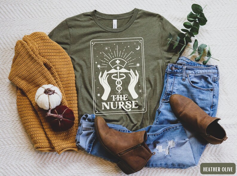 May include: A green t-shirt featuring a white graphic of a tarot card. The card depicts a white caduceus symbol with the text "The Nurse" underneath. The t-shirt is displayed on a white surface alongside a brown sweater, blue jeans, and brown boots.