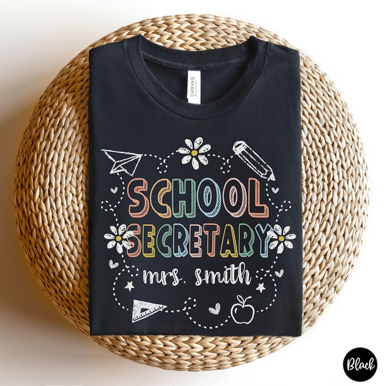 Peut inclure: T-shirt noir avec l'inscription "SCHOOL SECRETARY mrs. smith" en lettres color&eacute;es et soulign&eacute;es. Le motif comprend un avion en papier, un crayon, des fleurs et une pomme. Le t-shirt est pr&eacute;sent&eacute; sur une surface tiss&eacute;e.