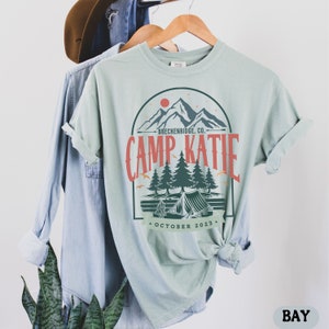May include: A light blue t-shirt with a vintage-style graphic design featuring a mountain range, trees, a tent, and the text "Camp Katie - Breckenridge, Co. - October 2023."