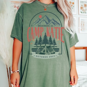 May include: A green oversized t-shirt with a vintage-inspired graphic design. The design features a mountain range, pine trees, a tent, and the text "Camp Katie - Breckenridge, Co. - October 2023".