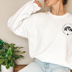 Peut inclure: Sweat-shirt blanc avec un graphique noir et blanc d'un symbole m&eacute;dical et le texte "Emergency Department RN" dans un cercle.