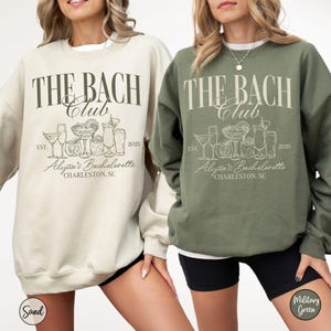 K&ouml;nnte beinhalten: Zwei Sweatshirts in Sand und Milit&auml;rgr&uuml;n mit dem Text "The Bach Club" und einer Grafik von Cocktailgl&auml;sern. Der Text "Alyssa's Bachelorette" und "Charleston, SC" sind ebenfalls auf den Sweatshirts enthalten.