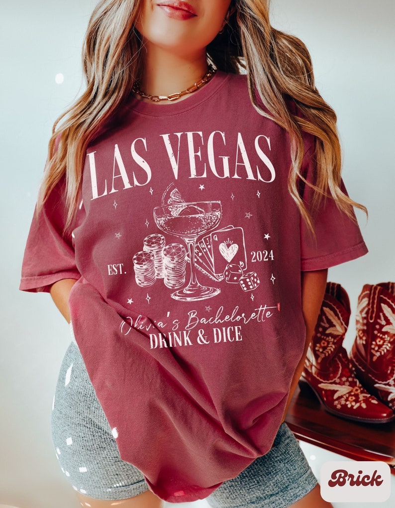 May include: A maroon t-shirt with a white graphic design featuring a cocktail glass, playing cards, dice, and the text "Las Vegas Est. 2024 Viva's Bachelorette Drink & Dice".