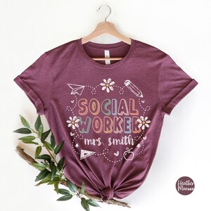 May include: Heather maroon t-shirt with the words "SOCIAL WORKER" in colorful letters, surrounded by daisies, a pencil, a paper airplane, and an apple. The text "mrs. smith" is below the words "SOCIAL WORKER".