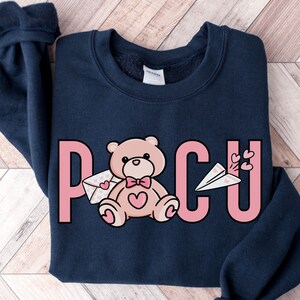 Peut inclure: Un sweat-shirt bleu marine avec un motif graphique rose qui dit "PCU" avec un ours en peluche tenant une lettre et un avion en papier avec des c&oelig;urs qui s'envolent.