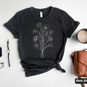 K&ouml;nnte beinhalten: Dunkelgraues Heather-T-Shirt mit einer wei&szlig;en Linienzeichnung eines Blumenstrau&szlig;es. Das Shirt liegt flach auf einer wei&szlig;en Oberfl&auml;che mit einer Uhr, einer Brille, einer kleinen Schmuckschale, einer braunen Ledertasche und einer Tasse Kaffee.