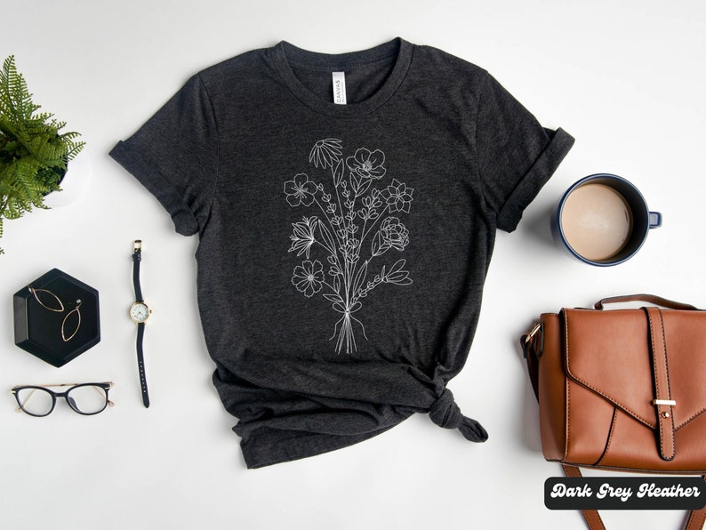 K&ouml;nnte beinhalten: Dunkelgraues Heather-T-Shirt mit einer wei&szlig;en Linienzeichnung eines Blumenstrau&szlig;es. Das Shirt liegt flach auf einer wei&szlig;en Oberfl&auml;che mit einer Uhr, einer Brille, einer kleinen Schmuckschale, einer braunen Ledertasche und einer Tasse Kaffee.