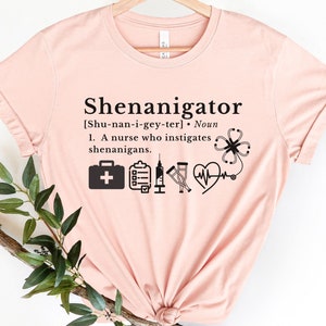 May include: A pink t-shirt with the text "Shenanigator" and a definition of the word. The definition includes the phonetic spelling of the word and a description of a nurse who instigates shenanigans. The shirt also features several medical icons, including a first aid kit, a clipboard, crutches, a stethoscope, and a heart.