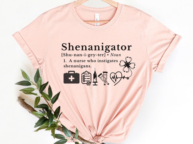May include: A pink t-shirt with the text "Shenanigator" and a definition of the word. The definition includes the phonetic spelling of the word and a description of a nurse who instigates shenanigans. The shirt also features several medical icons, including a first aid kit, a clipboard, crutches, a stethoscope, and a heart.