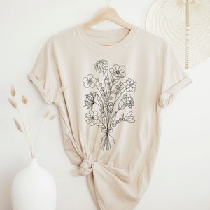 K&ouml;nnte beinhalten: Ein beiges T-Shirt mit einer schwarzen Linienzeichnung eines Blumenstrau&szlig;es. Das Shirt ist aus einem weichen, bequemen Material gefertigt und hat eine lockere Passform.