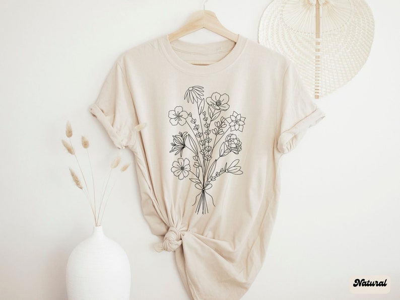 K&ouml;nnte beinhalten: Ein beiges T-Shirt mit einer schwarzen Linienzeichnung eines Blumenstrau&szlig;es. Das Shirt ist aus einem weichen, bequemen Material gefertigt und hat eine lockere Passform.