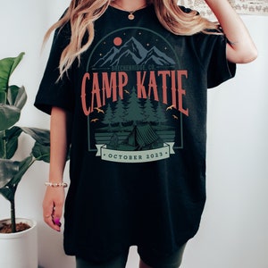 May include: Black oversized t-shirt with a graphic design of a mountain range, a tent, and trees. The text "Breckenridge Co. Camp Katie October 2023" is printed on the shirt.