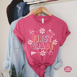 May include: A magenta t-shirt with the words "FIRST GRADE" in colorful block letters, surrounded by white daisies and school-themed illustrations. The shirt is hanging on a wooden hanger with a denim shirt and jeans.