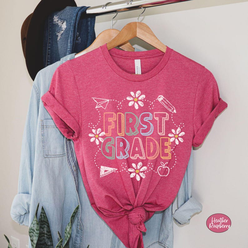 May include: A magenta t-shirt with the words "FIRST GRADE" in colorful block letters, surrounded by white daisies and school-themed illustrations. The shirt is hanging on a wooden hanger with a denim shirt and jeans.