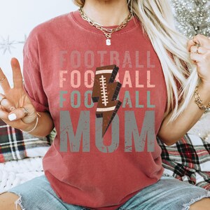 Op de afbeelding: Rood T-shirt met een verweerd grafisch ontwerp met de tekst "Football Mom" met een voetbal- en bliksemgrafiek.