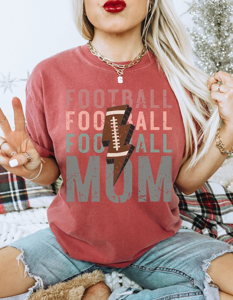 Op de afbeelding: Rood T-shirt met een verweerd grafisch ontwerp met de tekst "Football Mom" met een voetbal- en bliksemgrafiek.